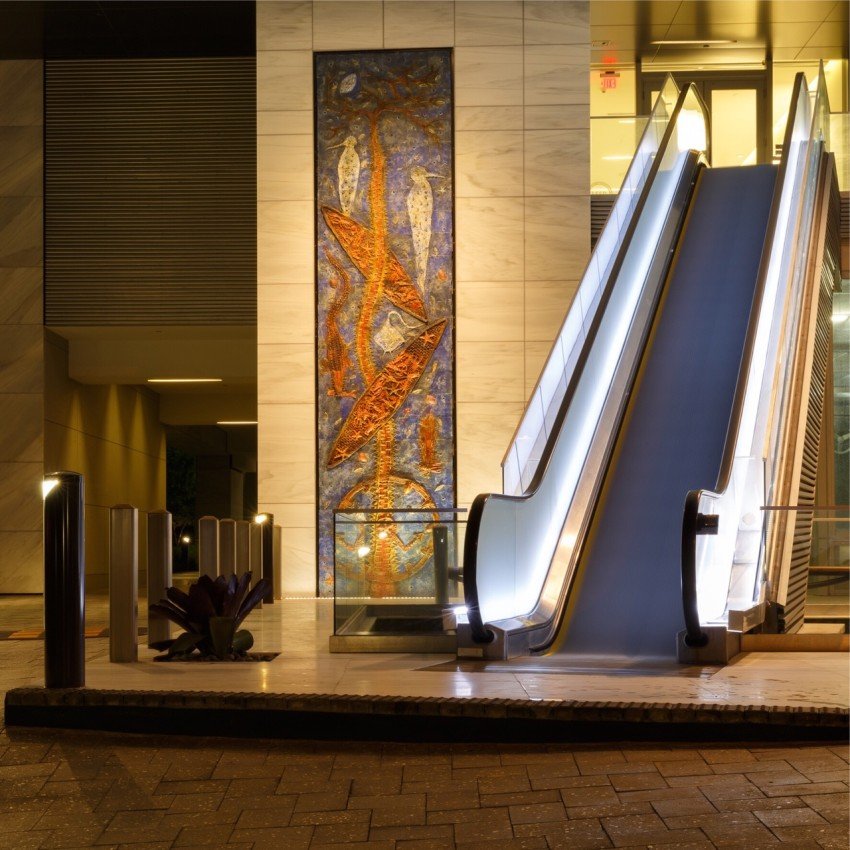 Lobby entrance of a commercial building in Aventura at night, with an illuminated mosaic art panel, washed marble walls and lit escalators